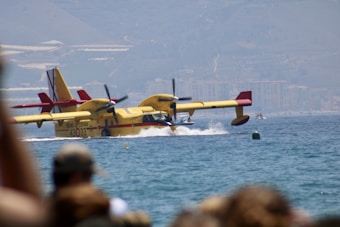A yellow amphibious firefighting aircraft with red accents is landing or taking off on water. Its propellers are spinning, and a splash of water is visible beneath it. In the background, a cityscape with buildings can be seen in the distance. People in the foreground are observing the scene.