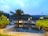 A tranquil waterside view with modern bungalows set among lush greenery. A palm tree and potted plants adorn the foreground, with the calm water reflecting the overcast sky and buildings. The setting suggests a peaceful and picturesque vacation spot.