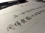 A close-up view of handwritten text in black ink on lined paper, with a pen placed at the top. The focus is on the text, while the pen and background are slightly blurred.