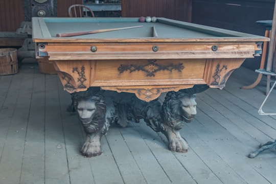 An intricately carved antique billiard table is adorned with lion sculptures as legs. The table is situated in a rustic room with wooden floors and various wooden furniture items in the background. A billiard cue and some balls rest on the table.