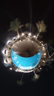 A spherical panoramic view of an illuminated poolside at night with palm trees surrounding a glowing blue swimming pool. The bright lights cast reflections on the water, creating a serene and mystical atmosphere. The perspective warps the pool area into a circular shape, resembling a small planet.