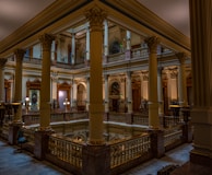 An elegant interior featuring grand architectural elements such as tall, ornate columns and intricately designed railings. The lighting is warm, highlighting the detailed moldings and the stained glass window. The setting appears to be a historic or government building with a sophisticated and classic design.