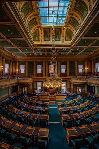A large and ornate legislative chamber with rows of wooden desks and dark green chairs arranged in a semicircle. The room features a high, vaulted ceiling with intricate designs and a central skylight. A grand chandelier hangs prominently from the ceiling. Stained glass windows and decorative panels adorn the walls, and a raised podium is situated at the front of the room.