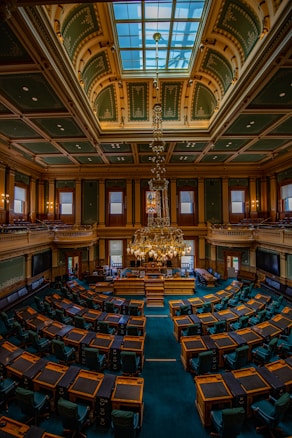 A large and ornate legislative chamber with rows of wooden desks and dark green chairs arranged in a semicircle. The room features a high, vaulted ceiling with intricate designs and a central skylight. A grand chandelier hangs prominently from the ceiling. Stained glass windows and decorative panels adorn the walls, and a raised podium is situated at the front of the room.