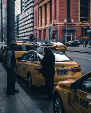A busy urban street scene featuring several yellow taxis lined up along the roadside. A person stands outside one of the taxis engaging with the driver. The street is flanked by tall buildings with prominent architecture, with a few scattered pedestrians and vehicles passing by.