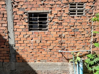A brick wall with uneven and mismatched mortar, featuring a small barred window on the upper left-half and two metal ventilation grates on the upper right. A gray concrete beam borders the left side, and a few green plants grow near the base, where blue pipes are also visible.