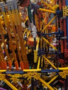 An intricate and colorful structure made from interlocking plastic rods and connectors, resembling a complex geometric model. The assembly consists of various hues including yellow, orange, and blue, suggesting a playful and creative design.