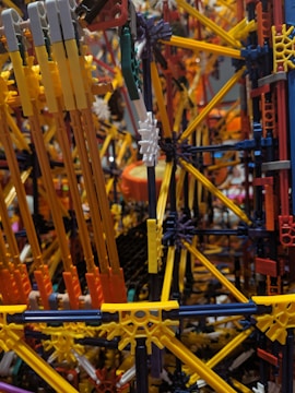 An intricate and colorful structure made from interlocking plastic rods and connectors, resembling a complex geometric model. The assembly consists of various hues including yellow, orange, and blue, suggesting a playful and creative design.