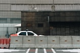A partially visible police car is parked behind concrete barriers in front of a modern, industrial-looking building. The building facade is composed of dark stone and metal grills, and there is a sense of security with additional fencing and barbed wire on top.