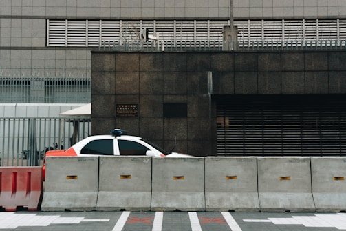 A partially visible police car is parked behind concrete barriers in front of a modern, industrial-looking building. The building facade is composed of dark stone and metal grills, and there is a sense of security with additional fencing and barbed wire on top.