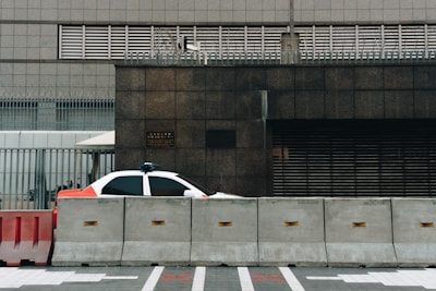A partially visible police car is parked behind concrete barriers in front of a modern, industrial-looking building. The building facade is composed of dark stone and metal grills, and there is a sense of security with additional fencing and barbed wire on top.
