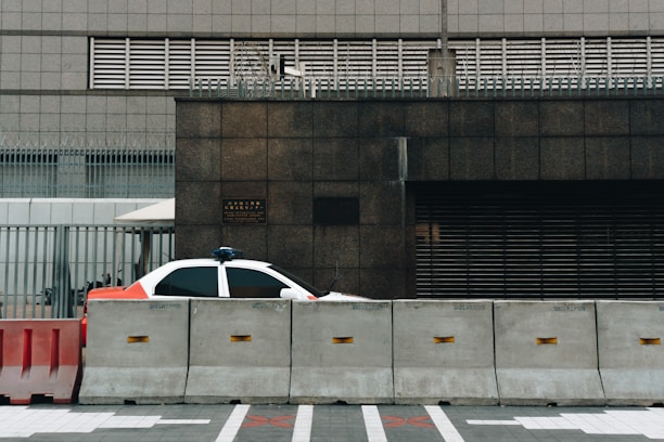 A partially visible police car is parked behind concrete barriers in front of a modern, industrial-looking building. The building facade is composed of dark stone and metal grills, and there is a sense of security with additional fencing and barbed wire on top.