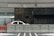 A partially visible police car is parked behind concrete barriers in front of a modern, industrial-looking building. The building facade is composed of dark stone and metal grills, and there is a sense of security with additional fencing and barbed wire on top.