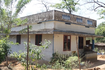 A one-story building with a flat roof, featuring large rectangular windows with dark red frames. The walls are painted white, and there is an overhang above the entrance. Surrounding the house are various plants and trees, indicating a rural setting. The structure shows signs of weathering with some discoloration on the exterior.