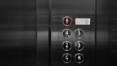 A set of elevator buttons with numbers for floors, including a highlighted button with the number 6 lit in red, indicating the selected floor. Other buttons visible are labeled with numbers and letters, likely representing different floors and perhaps a ground or main floor.