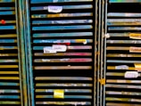 Several storage racks with a variety of colored slats, which appear to be used for organizing or holding paintings or paper. Labels are attached to some of the slats, suggesting organization or categorization. The racks are metallic with peeling paint, indicating frequent use or age.