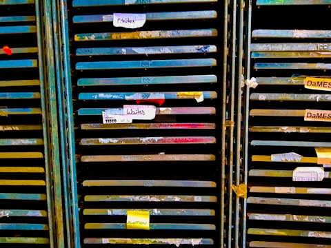 Several storage racks with a variety of colored slats, which appear to be used for organizing or holding paintings or paper. Labels are attached to some of the slats, suggesting organization or categorization. The racks are metallic with peeling paint, indicating frequent use or age.