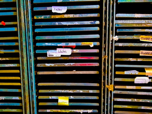 Several storage racks with a variety of colored slats, which appear to be used for organizing or holding paintings or paper. Labels are attached to some of the slats, suggesting organization or categorization. The racks are metallic with peeling paint, indicating frequent use or age.