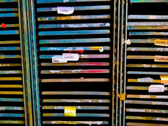 Several storage racks with a variety of colored slats, which appear to be used for organizing or holding paintings or paper. Labels are attached to some of the slats, suggesting organization or categorization. The racks are metallic with peeling paint, indicating frequent use or age.