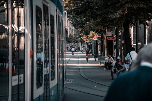 A bustling urban street lined with trees and benches. People are engaged in various activities: walking, sitting on benches, and cycling down the pedestrian walkway. A tram is visible on the left side, reflecting its surroundings. Signs and buildings add to the urban atmosphere, and the dappled sunlight creates a warm, inviting ambiance.