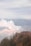A volcano emitting thick white smoke surrounded by rugged landscapes and sparse vegetation. In the foreground, a group of people stands on a viewing platform, observing the scene. The overcast sky casts a muted light over the entire view, creating a serene yet somewhat dramatic atmosphere.