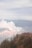 A volcano emitting thick white smoke surrounded by rugged landscapes and sparse vegetation. In the foreground, a group of people stands on a viewing platform, observing the scene. The overcast sky casts a muted light over the entire view, creating a serene yet somewhat dramatic atmosphere.