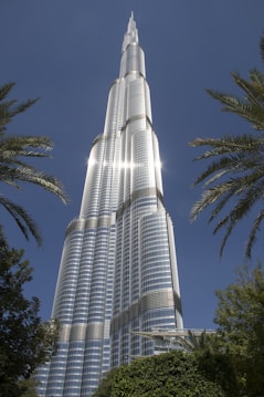 A towering skyscraper with a sleek, modern design reaches towards a clear blue sky. The structure, made primarily of glass and steel, reflects the sunlight brilliantly. Palm trees frame the image on both sides, adding a touch of nature to the urban scene.
