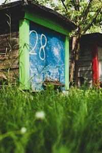 An old wooden beehive painted with bright blue, green, and red sections is surrounded by lush green grass. The number 28 is prominently painted on the front panel. The setting appears to be a garden or an orchard, with trees and plants adding natural beauty to the surroundings.