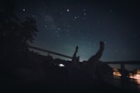 A person is lying down on a deck or platform at night, gazing up at a clear starry sky. Silhouetted trees and a calm body of water in the foreground are faintly illuminated by lights, while the vastness of the universe is visible overhead.