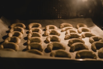 A tray of crescent-shaped pastries is arranged on parchment paper, baking in an oven. The warm light casts soft shadows, indicating they are in the process of rising and baking.