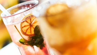 A close-up of two refreshing beverages, one is pink with a visible slice of lemon and mint leaves, and the other is a blurred orange drink. Both are served in clear glasses with ice cubes visible.