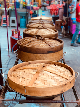 A row of large, round bamboo steamers with woven lids placed on a metal rack, likely in a street market setting. The background includes vibrant market scenes with people and colorful objects.
