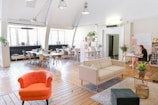 A spacious, modern office area featuring wooden floors and large windows allowing natural light. A cozy furniture arrangement includes a red armchair, a beige sofa, and a small table. Several potted plants are placed throughout the space, adding greenery. A large table with chairs is arranged near the windows, suitable for meetings. A woman is working on a laptop at a desk on the right side.