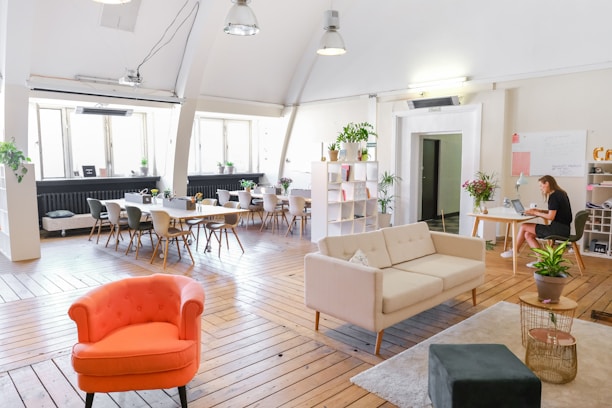 A spacious, modern office area featuring wooden floors and large windows allowing natural light. A cozy furniture arrangement includes a red armchair, a beige sofa, and a small table. Several potted plants are placed throughout the space, adding greenery. A large table with chairs is arranged near the windows, suitable for meetings. A woman is working on a laptop at a desk on the right side.