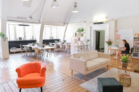 A spacious, modern office area featuring wooden floors and large windows allowing natural light. A cozy furniture arrangement includes a red armchair, a beige sofa, and a small table. Several potted plants are placed throughout the space, adding greenery. A large table with chairs is arranged near the windows, suitable for meetings. A woman is working on a laptop at a desk on the right side.
