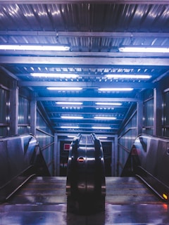 An indoor escalator leads downward, surrounded by metal walls and ceiling panels. The setting is illuminated by bright fluorescent lights, creating a futuristic and industrial atmosphere. The shiny surfaces reflect the lights, enhancing the modern aesthetic.
