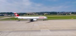 An airplane bearing the 'SWISS' logo is taxiing on a runway with green fields and hills in the background. The aircraft is predominantly white with a red tail fin featuring a white cross. The sky is clear and blue with a few scattered clouds.