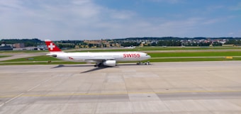 An airplane bearing the 'SWISS' logo is taxiing on a runway with green fields and hills in the background. The aircraft is predominantly white with a red tail fin featuring a white cross. The sky is clear and blue with a few scattered clouds.