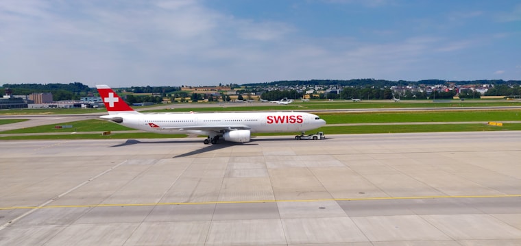An airplane bearing the 'SWISS' logo is taxiing on a runway with green fields and hills in the background. The aircraft is predominantly white with a red tail fin featuring a white cross. The sky is clear and blue with a few scattered clouds.