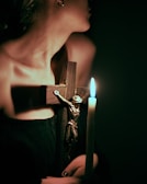 A person is holding a wooden crucifix featuring a metallic figure of Christ, along with a lit candle casting a soft glow in the dimly lit environment. The image conveys a sense of spirituality or religious contemplation.