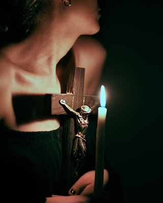 A person is holding a wooden crucifix featuring a metallic figure of Christ, along with a lit candle casting a soft glow in the dimly lit environment. The image conveys a sense of spirituality or religious contemplation.