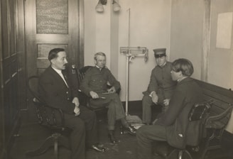 Four men are seated in a room that appears to be an office or waiting area, with wooden furniture and a scale visible in the background. Three of the men are dressed in uniforms, while one wears a suit. The men are engaged in conversation or a meeting, sitting in chairs and on a bench along the wall.