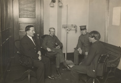 Four men are seated in a room that appears to be an office or waiting area, with wooden furniture and a scale visible in the background. Three of the men are dressed in uniforms, while one wears a suit. The men are engaged in conversation or a meeting, sitting in chairs and on a bench along the wall.