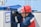 A person wearing a red hard hat and a protective face shield is working on an electrical or utility box. The individual is dressed in a navy blue and red uniform. A sign in Russian is hanging on the box, reading 'Work Here'. The background is slightly blurred, with hints of industrial elements.
