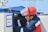 A person wearing a red hard hat and a protective face shield is working on an electrical or utility box. The individual is dressed in a navy blue and red uniform. A sign in Russian is hanging on the box, reading 'Work Here'. The background is slightly blurred, with hints of industrial elements.