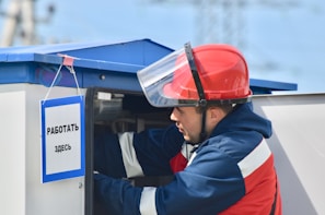 A person wearing a red hard hat and a protective face shield is working on an electrical or utility box. The individual is dressed in a navy blue and red uniform. A sign in Russian is hanging on the box, reading 'Work Here'. The background is slightly blurred, with hints of industrial elements.