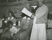 An older person is standing while reading a book or document to a group of seated people. The seated individuals, including a child, appear to be listening intently. The setting seems to be indoors, possibly a wooden cabin or similar structure.