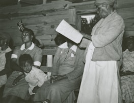 An older person is standing while reading a book or document to a group of seated people. The seated individuals, including a child, appear to be listening intently. The setting seems to be indoors, possibly a wooden cabin or similar structure.