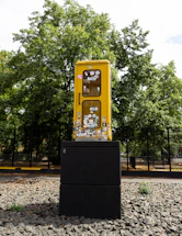 A yellow phone booth is mounted on a black pedestal in an outdoor setting. The booth is covered with various colorful stickers and is surrounded by a gravel surface. Tall green trees are visible in the background, and a fence can be seen lining the perimeter.