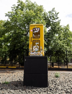 A yellow phone booth is mounted on a black pedestal in an outdoor setting. The booth is covered with various colorful stickers and is surrounded by a gravel surface. Tall green trees are visible in the background, and a fence can be seen lining the perimeter.